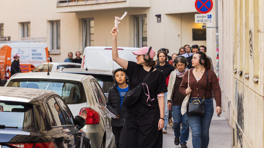 Gruppe von Menschen mit Kopfhörern geht auf einem Gehweg zwischen geparkten Autos und Gebäuden, eine Person hält ein weißes Objekt in die Luft
