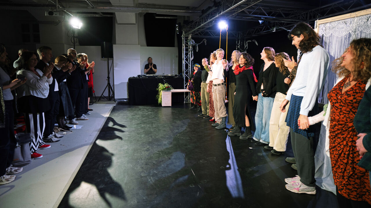 Menschen stehen in zwei Reihen einander gegenüber in einem Theaterraum und klatschen, eine Person in der Mitte hebt die Hand