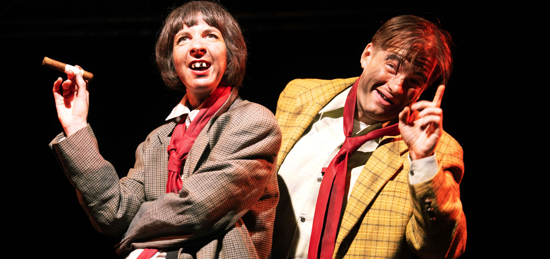 Zwei Theaterschauspieler, eine Frau und ein Mann, stehen auf der Bühne. Beide sind absichtlich verwahrlost gekleidet, die Frau hält eine Zigarre in der Hand, beide tragen Sakkos und rote Krawatten, ihre Gesichter sind mit Dreck beschmiert, die Frau hat mehrere schwarzgefärbte Zähne.