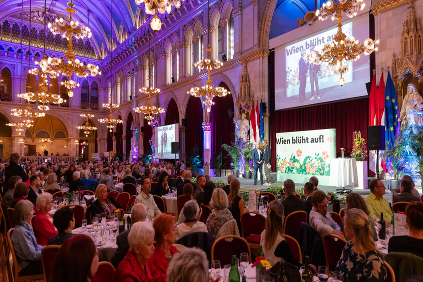 Blick in den vollbesetzten großen Festsaal des Wiener Rathauses. An den Tischen sitzen Menschen, im Vordergrund eine Bühne auf der gerade jemand von Karl Ploberger interviewt wird. Alles ist prächtig mit Blumen in den unterschildichsten Farben geschmückt.