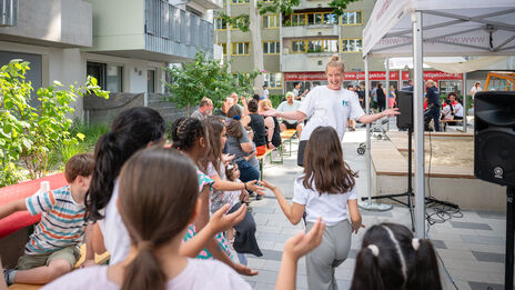 Im Innenhof einer Wohnhausanlage tanzt eine Frau mit den anwesenden Kindern.