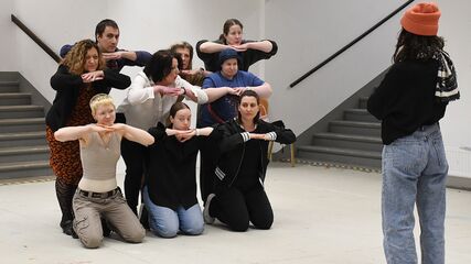 Drei Personen knien auf dem Boden und stützen ihre Köpfe auf den Händen ab. Die Arme liegen dabei imaginär auf einer Mauer oder einem Tisch. Dahinter stehen drei weitere PErsonen nach vorne gebeugt und tun es ihnen gleich. Dahinter stehen weitere drei PErsonen mit der selben Geste. Vor dieser Gruppe steht eine Person mit dem Rücken zur Kamera gewandt.