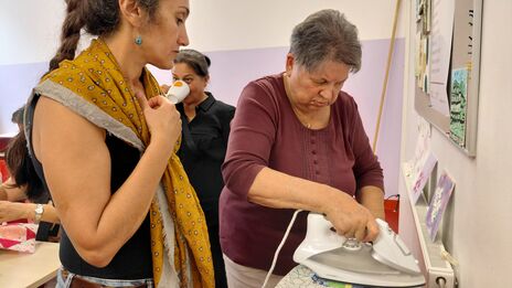 Ältere Frau bügelt Kleidung auf einem Tisch, während eine jüngere Frau mit gelbem Schal daneben steht und zuschaut