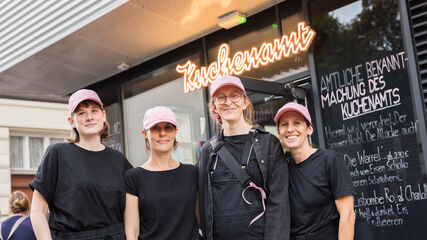 Vier Frauen in schwarzer Arbeitskleidung und rosa Kappen stehen vor einem Gebäude mit einem leuchtenden Schriftzug 'Kuchenamt' und einer Tafel mit handgeschriebener Speisekarte.