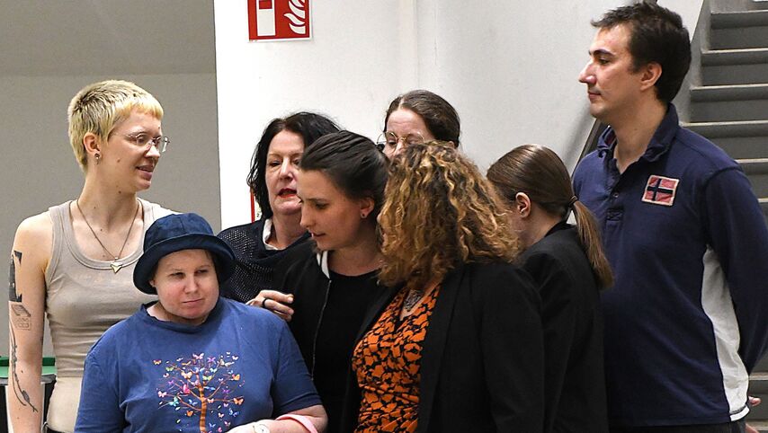 Eine Gruppe von Menschen steht dicht gedrängt beieinander in einem Raum mit Stiegen im Hintergrund. Sie sprechen miteinander.