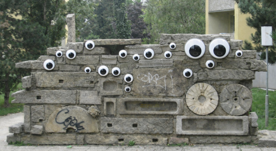 Betonskulptur in Mauerform mit mehreren aufgeklebten großen und kleinen runden Augen in einem Park