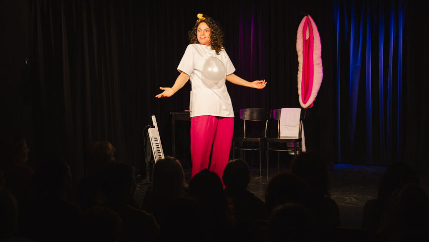 Eine Frau steht in einer pinken Hose und mit weißem T-Shirt, auf dem ein Luftballon in Höhe ihres Bauches hängt auf einer schwarz behangenen Bühne und macht eine fragende Geste mit den Armen. Neben ihr steht ein Umhängekeyboard (Keytar). Hinter erkennt man ein spezielles Bühnenkostüm, welches eine Vagina darstellen soll sowie zwei Stühle.
