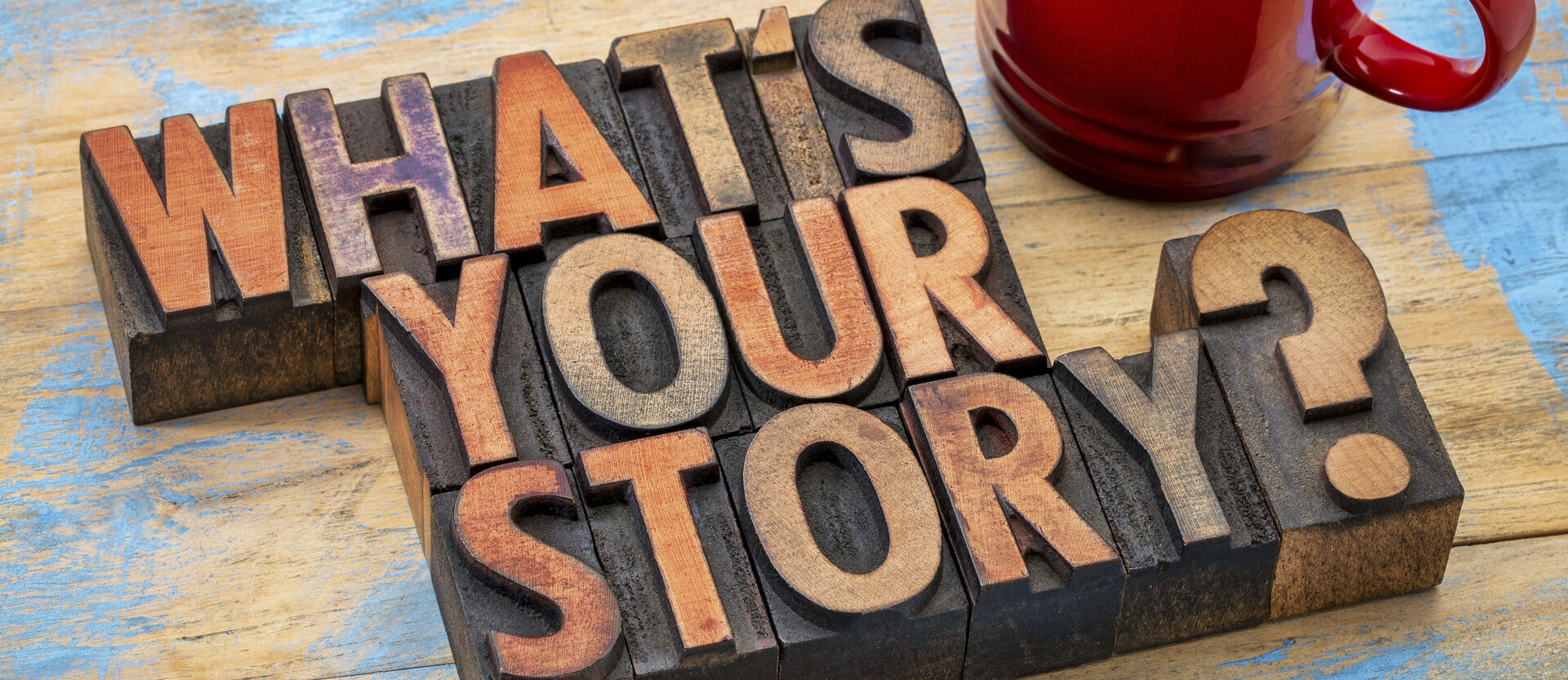 Ein Holzschild mit der Aufschrift "What's your story?" und eine rote Tasse mit Kaffee