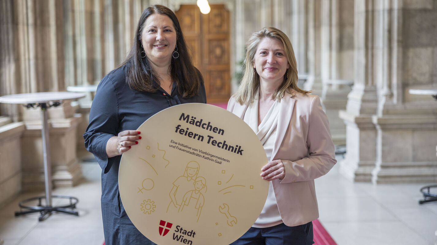 Vizebürgermeisterin und Frauenstadträtin Kathrin Gaál und Vizebürgermeisterin und Bildungsstadträtin Bettina Emmerling präsentieren die „Mädchen feiern Technik“-Workshops mit einem runden Schild, das von beiden gemeinsam gehaltne wird. Das Bild ist in den Arkadengängen des Rathauses aufgenommen worden. Kathrin Gaál trägt ein dunkelblaues Kleid, Bettina Emmerling eine dunkelblaue Hoseein beiges Oberteil mit Schalkragen und einen blassrosa Blazer darüber. Beide tragen ihre Haare offen.