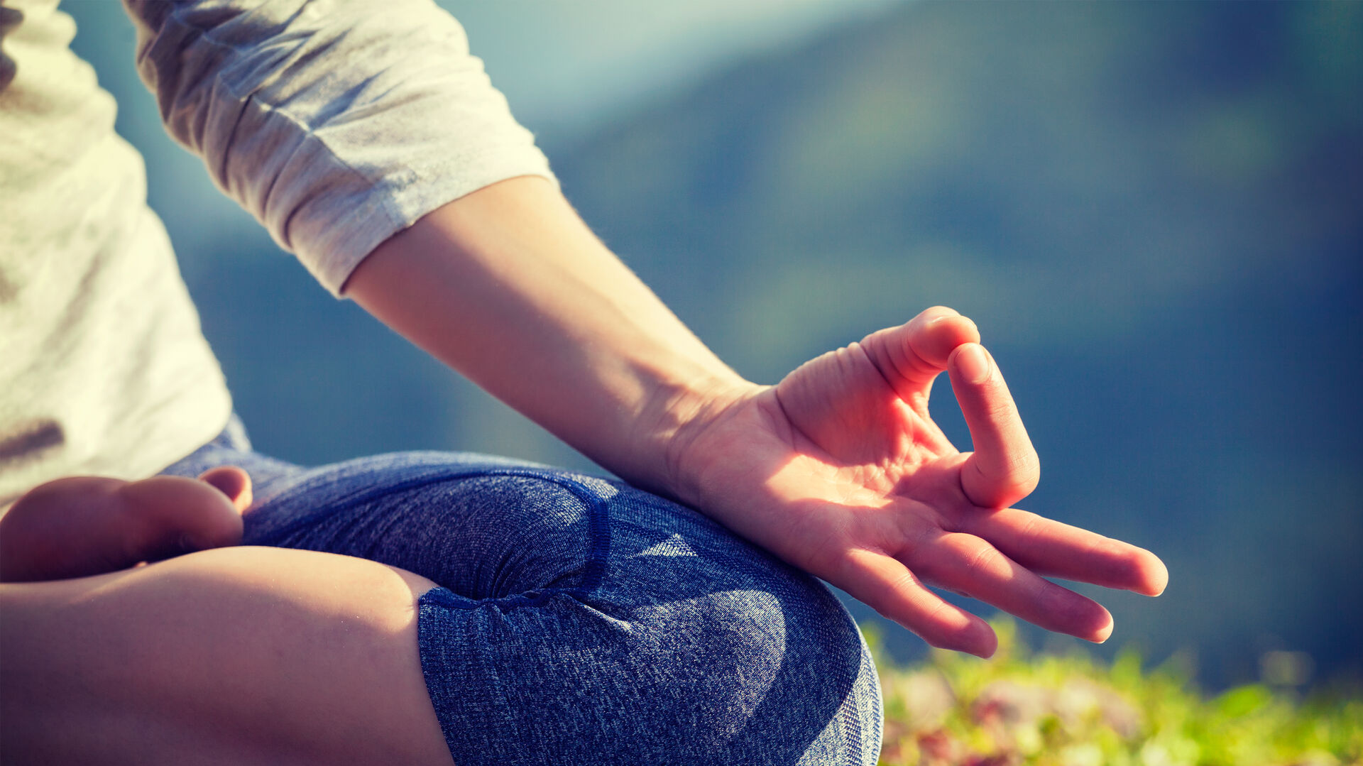 Nahaufnahme von Knie und Hand einer Person, die im Schneidersitz auf einer Gymnastikmatte sitzt und Daumen und Zeigefinger zu einem Mudra-Zeichen geformt hat.