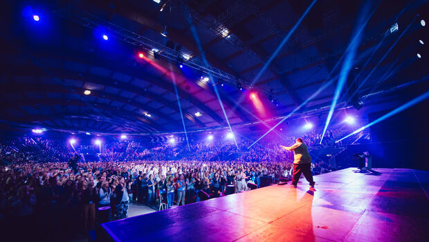 Person von hinten auf einer großen Bühne in einer riesigen Eventhalle, vollends gefüllt mit Menschen. 