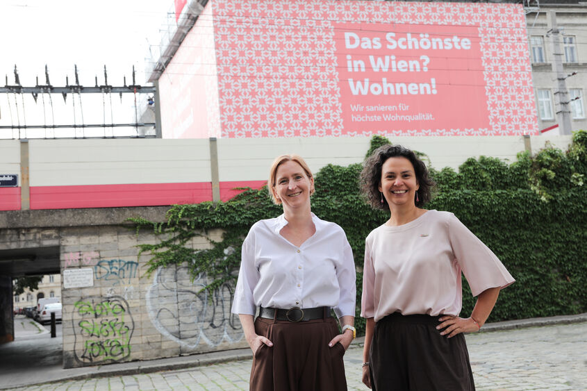 Zwei Frauen stehen vor einer mit Graffiti besprühten Mauer und einem Plakat mit der Aufschrift 'Das Schönste in Wien? Wohnen!'