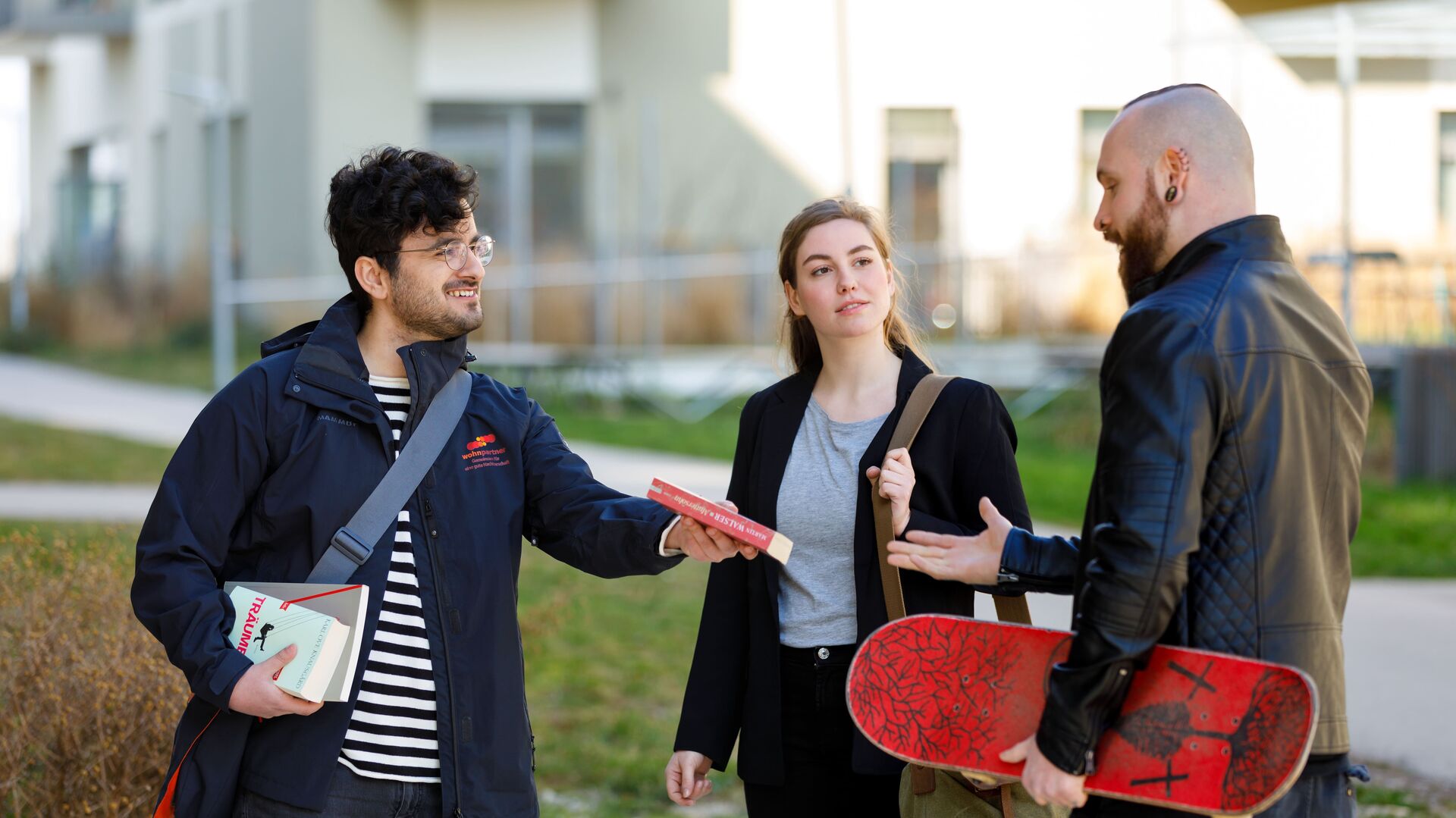 Ein wohnpartner-Mitarbeiter übergibt ein Buch an einen jungen Mann mit Skateboard unter dem Arm. Zwischen ihnen steht eine junge Frau mit einer Schultertasche.