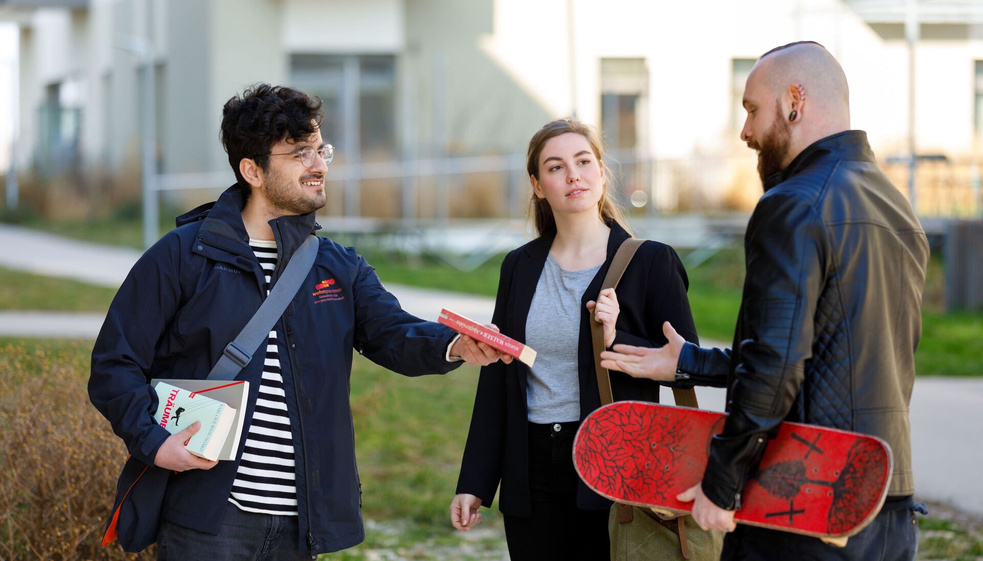 Ein wohnpartner-Mitarbeiter übergibt ein Buch an einen jungen Mann mit Skateboard unter dem Arm. Zwischen ihnen steht eine junge Frau mit einer Schultertasche.