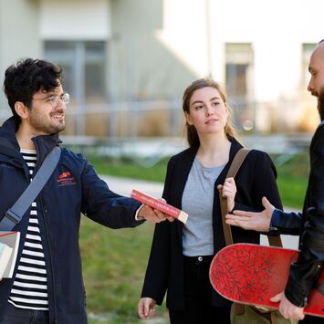 Ein wohnpartner-Mitarbeiter übergibt ein Buch an einen jungen Mann mit Skateboard unter dem Arm. Zwischen ihnen steht eine junge Frau mit einer Schultertasche.