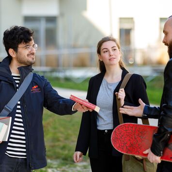 Ein wohnpartner-Mitarbeiter übergibt ein Buch an einen jungen Mann mit Skateboard unter dem Arm. Zwischen ihnen steht eine junge Frau mit einer Schultertasche.