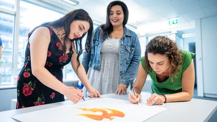 Drei Teilnehmerinnen der wohnpartner Akademie malen oder beschriften ein Plakat. in der Mitte des Plakats ist eine Art Feuervogel zu erkennen.