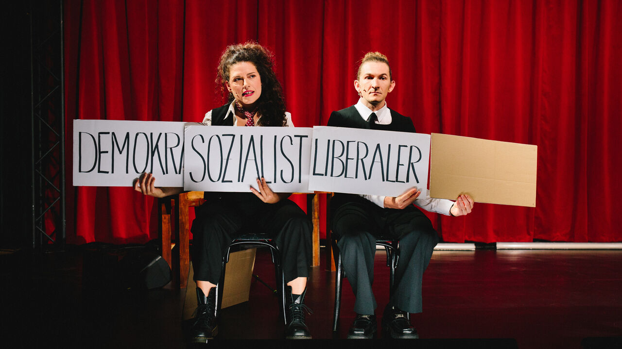 Zwei sitzende Personen vor rotem Vorhang halten jeweils ein Schild mit den Worten 'DEMOKR', 'SOZIALIST', 'LIBERALER' und einem leeren Schild