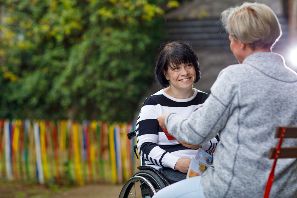 Beratungssituation im Freien, Rollstuhlfahrerin wird von einer Frau beraten, sie überreicht ihr einen Folder