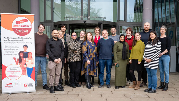Mehrere erwachsene Personen unterschiedlicher Ethnien stehen nebeneinander mit Blick zur Kamera. Neben Ihnen ist eine wohnpartner-Akademie-Beachflag platziert. 