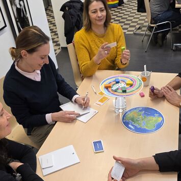 Fünf Personen sitzen um einen Tisch und spielen ein Brettspiel mit buntem Spielbrett und Karten