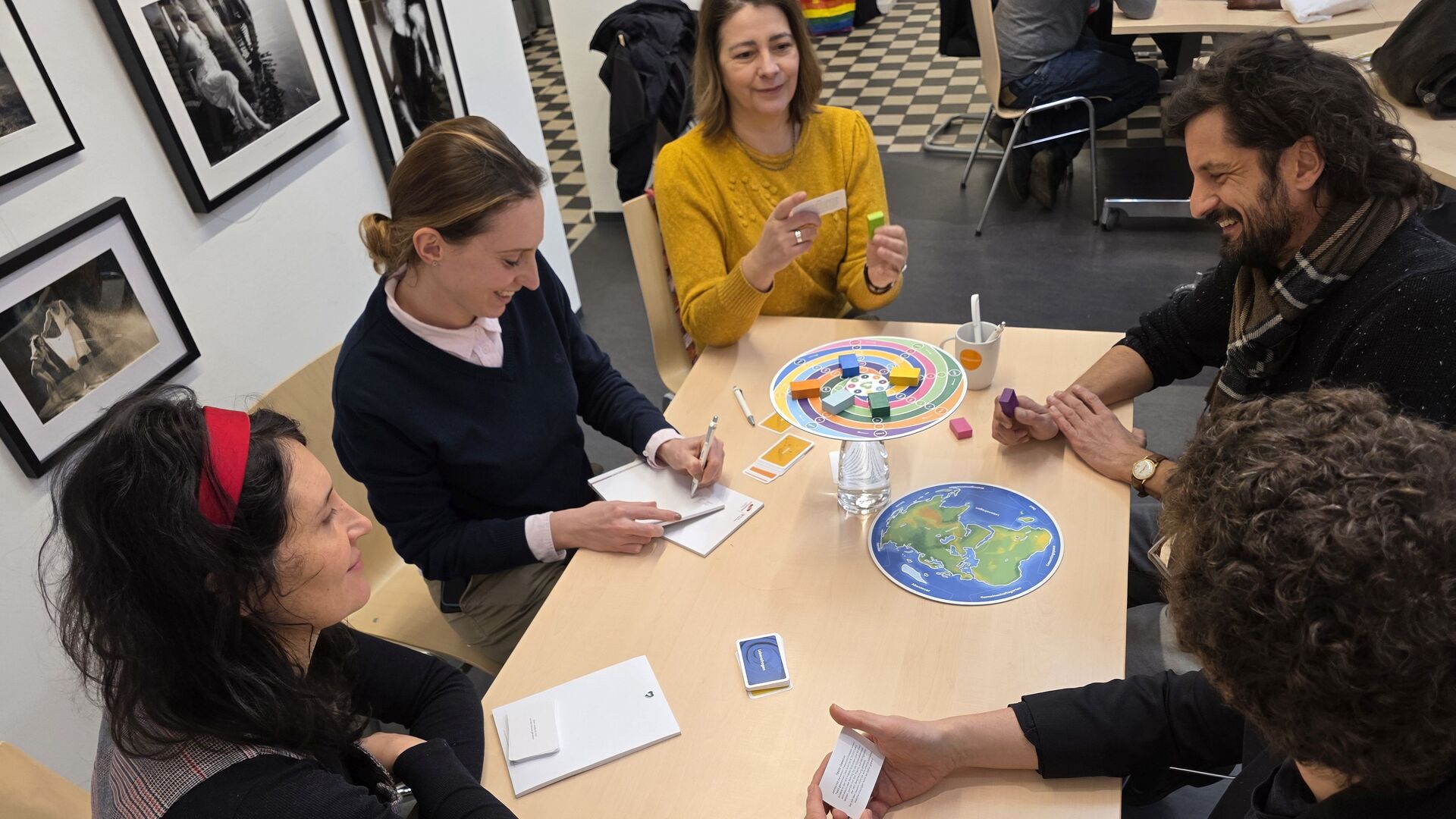 Fünf Personen sitzen um einen Tisch und spielen ein Brettspiel mit buntem Spielbrett und Karten