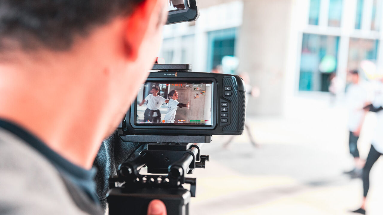 Person filmt tanzende Jugendliche mit einer Kamera im Freien; Szene ist auf dem Kameradisplay sichtbar.