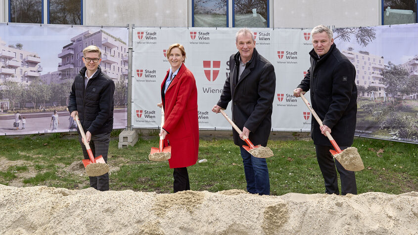 4 Personen stehen mit Spaten mit Sand darauf hinter einem Sandhaufen und lächeln in die Kamera. I Hintergrund ist ein Bauzaun mit Transparenten auf denen Visualisierungen des Gemeindebau NEU und das Logo von Stadt Wien/Wiener Wohnen zu erkennen ist.