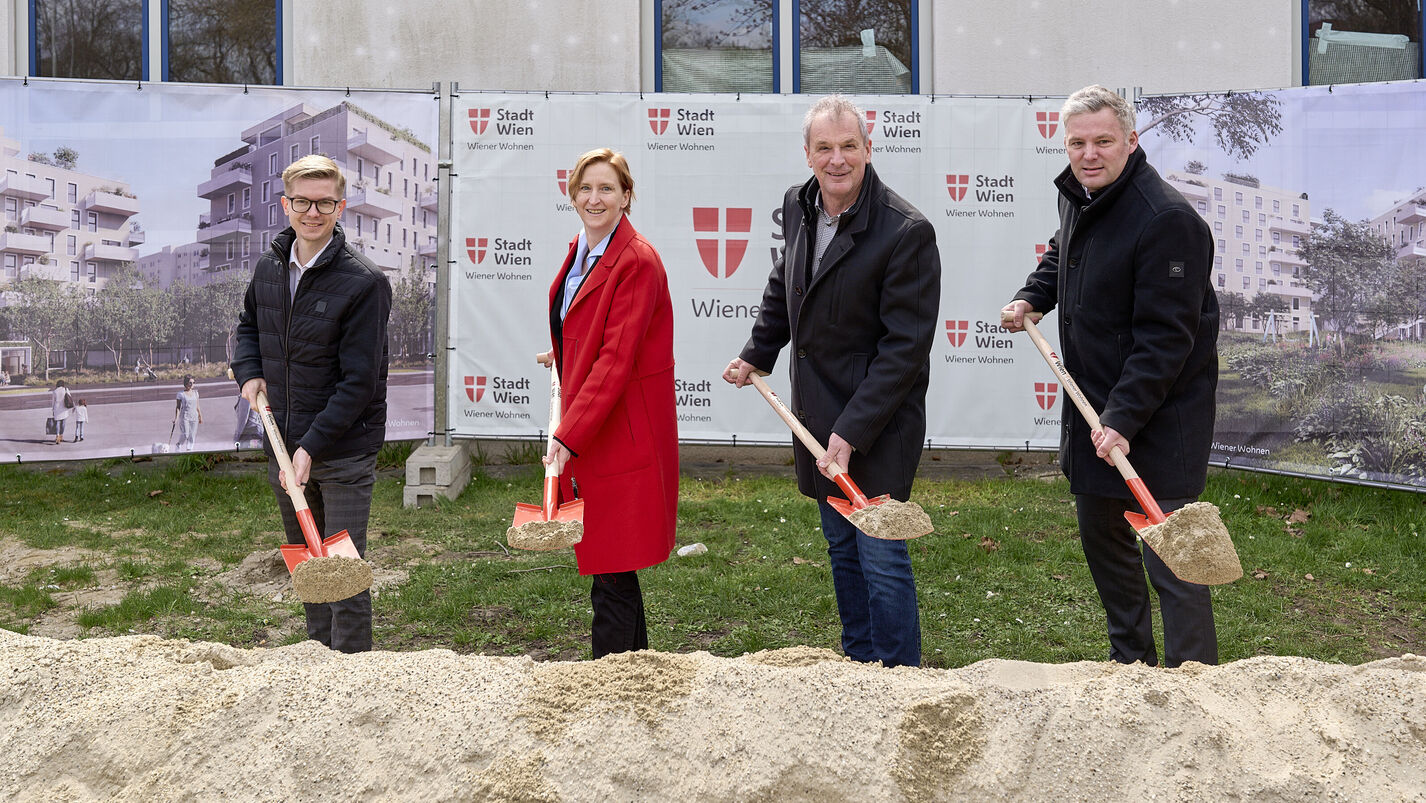 4 Personen stehen mit Spaten mit Sand darauf hinter einem Sandhaufen und lächeln in die Kamera. I Hintergrund ist ein Bauzaun mit Transparenten auf denen Visualisierungen des Gemeindebau NEU und das Logo von Stadt Wien/Wiener Wohnen zu erkennen ist.