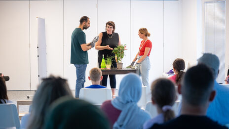 Drei Personen stehen an einem Tisch mit Pflanzen und sprechen vor einem sitzenden Publikum in einem hellen Raum mit Jalousien am Fenster.