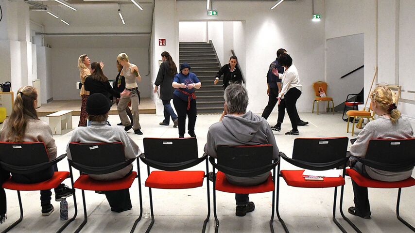 neun Personen tanzen jede für sich in einem Raum mit Stiegen im Hintergrund, Davor sitzen vier Personen und beobachten sie.