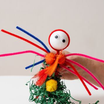 Bunte Bastelfigur mit Pfeifenreinigern, Pompons und Wackelaugen, gehalten von einer Hand vor hellem Hintergrund