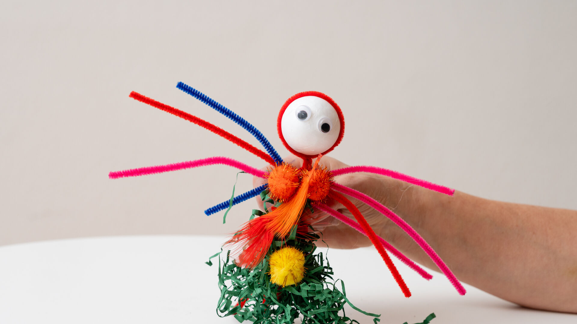 Bunte Bastelfigur mit Pfeifenreinigern, Pompons und Wackelaugen, gehalten von einer Hand vor hellem Hintergrund