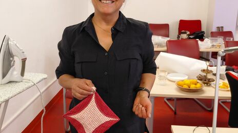 Lächelnde Frau in schwarzer Bluse hält einen rot-weiß gehäkelten Topflappen in einem Raum mit roten Stühlen und Tischen