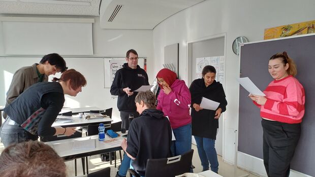 Mehrere Personen befinden sich in einem Klassenzimmer, Manche stehend, wenige sitzend. Sie sprechen miteinander und haben Zetteln in der Hand.