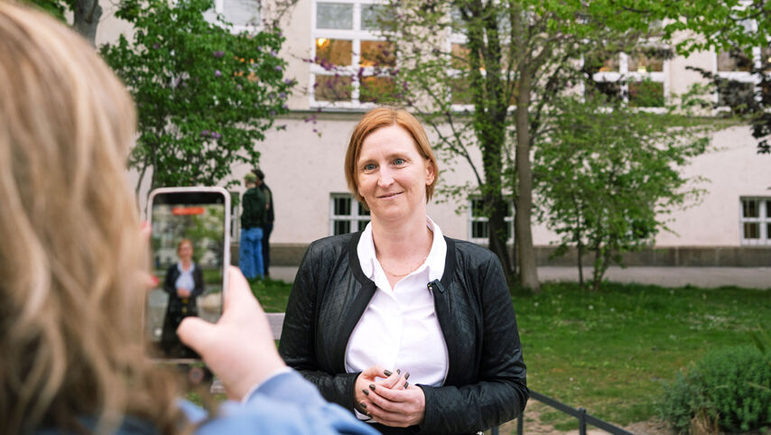 MAn blickt über die Schulter einer Person, die mit ihrem Handy ein Bild einer anderen Person macht, die vor einem Gebäude mit der Aufschrift Karl-Seitz-Hof steht.