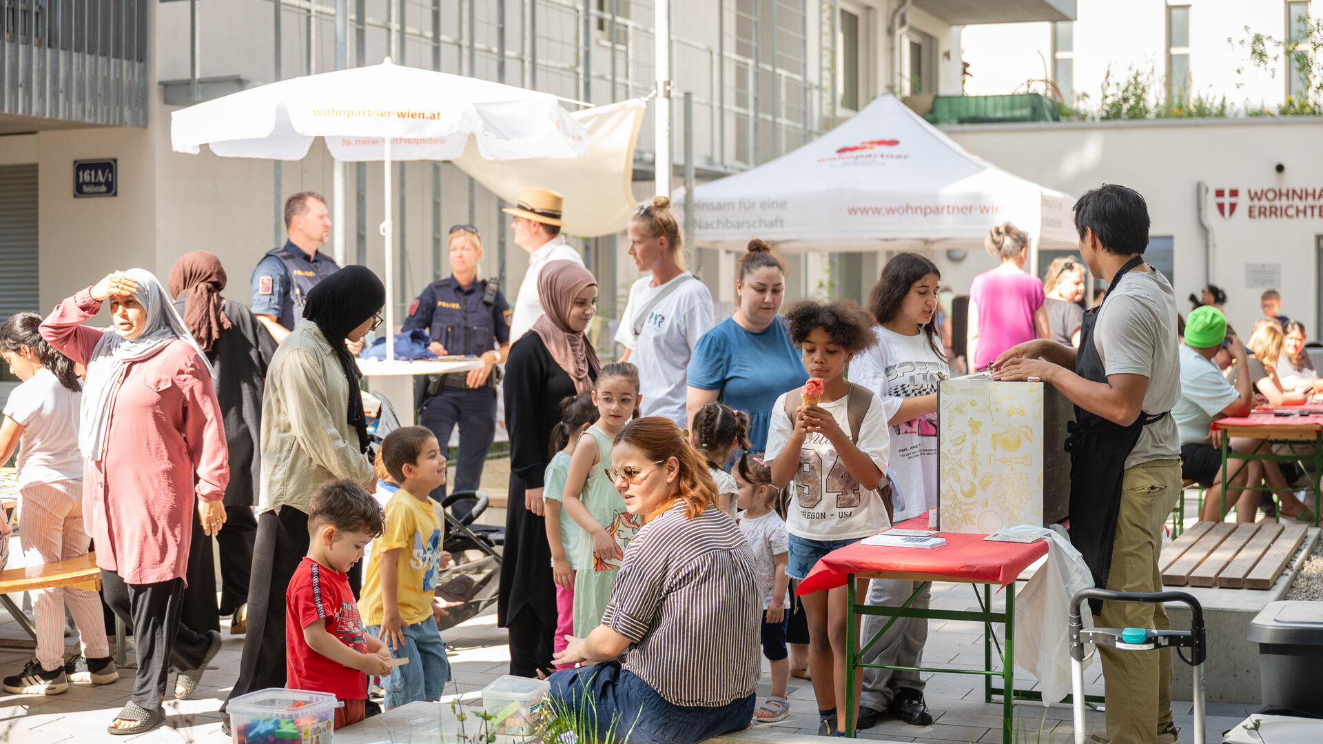 Menschen verschiedener Altersgruppen und Herkunft versammeln sich bei einem sonnigen Straßenfest mit Ständen und Sitzgelegenheiten in einem Innenhof