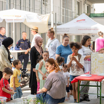 Menschen verschiedener Altersgruppen und Herkunft versammeln sich bei einem sonnigen Straßenfest mit Ständen und Sitzgelegenheiten in einem Innenhof