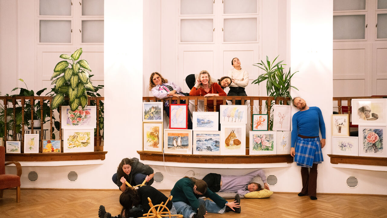 Gruppe von Menschen in einem hellen Innenraum eines Gemeindebaus, die gemeinsam eine performative Szene entwickeln; Zeichnungen und Pflanzen sind auf einer Balustrade arrangiert.