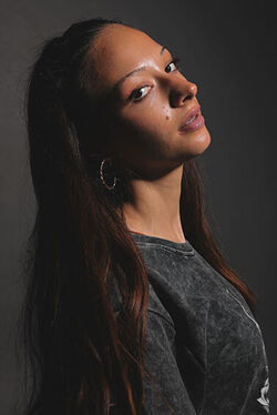 Person im Halbprofil. Sie ist gebräunt hat langes braunes Haar, einen Creolenohrring und hat den Mund leicht geöffnet. und Der Kopf ist leicht nach hinten geneigt, sie sieht in die Kamera