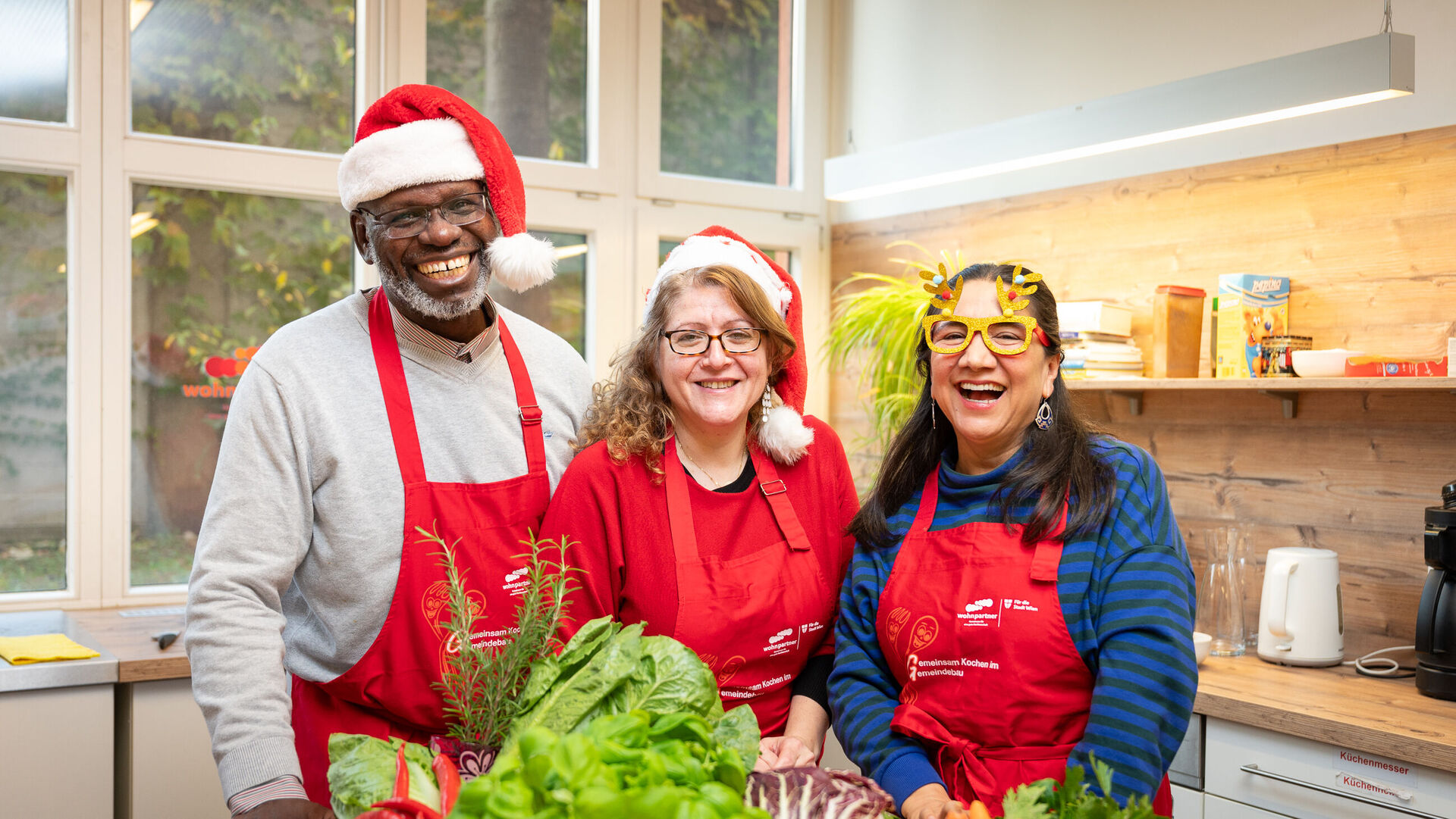 Drei Hobbyköch*innen stehen in einer wohnpartner-Küche und tragen Weihnachtsmützen und -brillen.