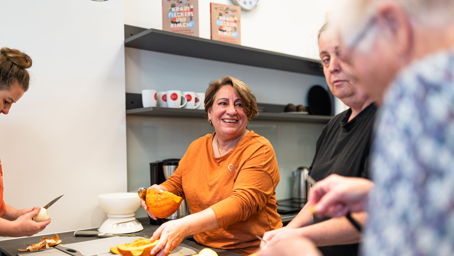 Mehrere Personen bereiten in einer Küche gemeinsam Kürbisse und anderes Gemüse zu, eine Frau in orangefarbenem Pullover lächelt dabei.