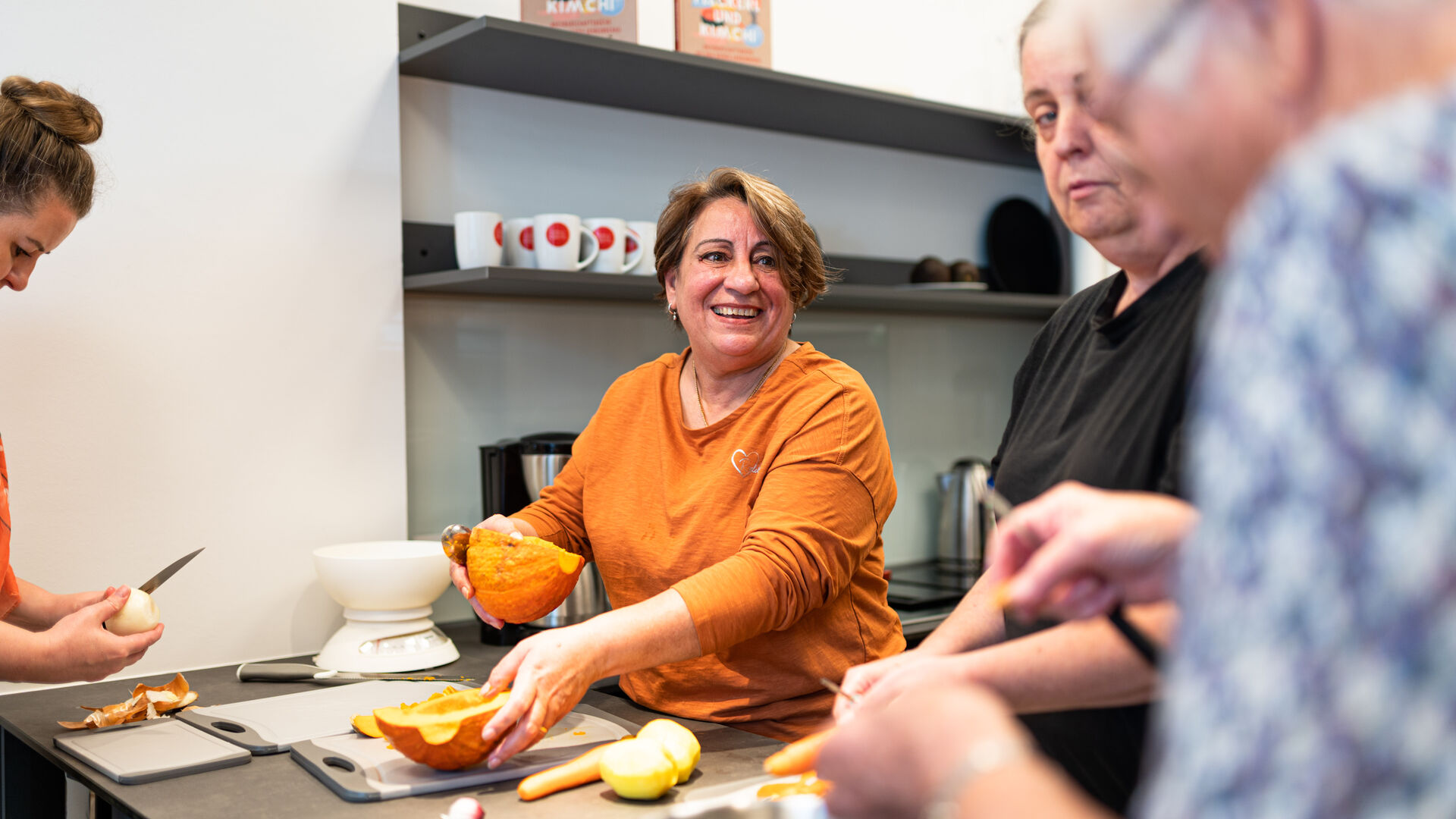 Mehrere Personen bereiten in einer Küche gemeinsam Kürbisse und anderes Gemüse zu, eine Frau in orangefarbenem Pullover lächelt dabei.