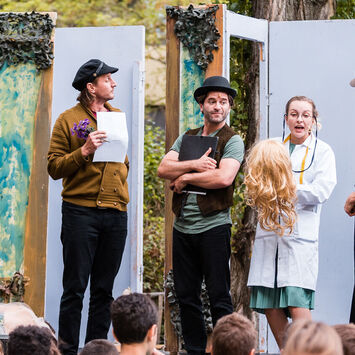 Theater-Schauspieler*innen treten auf einer Freiluftbühne vor Zuschauer*innen auf.