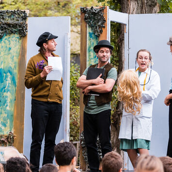 Theater-Schauspieler*innen treten auf einer Freiluftbühne vor Zuschauer*innen auf.