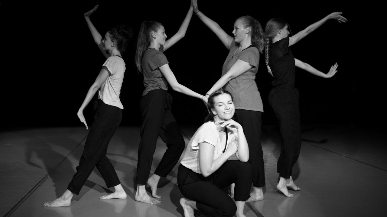 Fünf junge Frauen in einer Tanzpose auf einer Bühne, eine kniet vorne und lächelt, die anderen stehen dahinter mit erhobenen Armen.