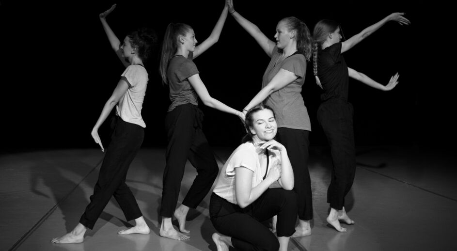 Fünf junge Frauen in einer Tanzpose auf einer Bühne, eine kniet vorne und lächelt, die anderen stehen dahinter mit erhobenen Armen.