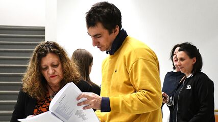 2 Personen stehen in einem Raum mit Stiegen im Hintergrund und lesen in einem Skript, im Hintergrund stehen weitere 3 Personen und unterhalten sich