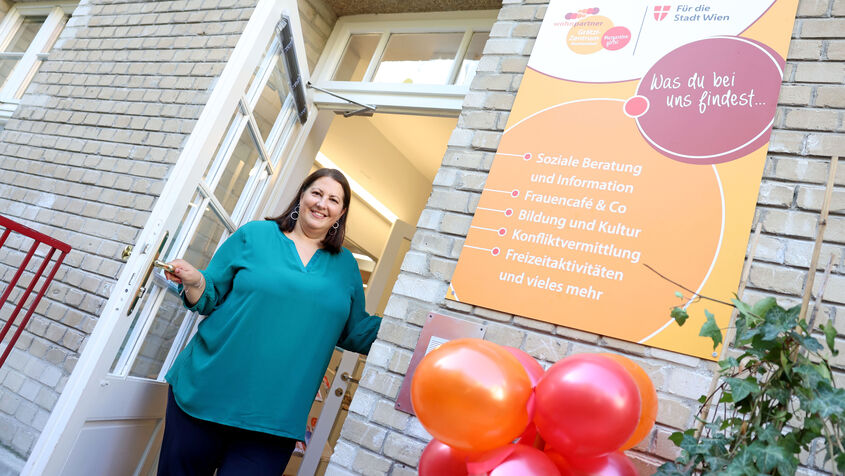 Wohnbaustadträtin Kathrin Gaál steht in der Türe eines wohnpartner Grätzl-Zentrums und lädt die Betrachter des Bildes dazu ein, herein zu kommen. Sie trägt eine Petrolfarbige Bluse und eine schwarze Hose.