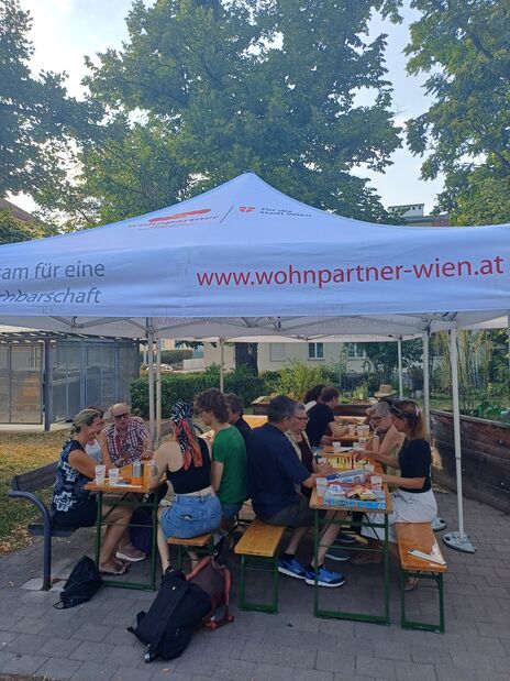 Menschen sitzen an zwei Holzbänken unter einem weißen Pavillon mit der Aufschrift 'www.wohnpartner-wien.at' und essen gemeinsam im Freien
